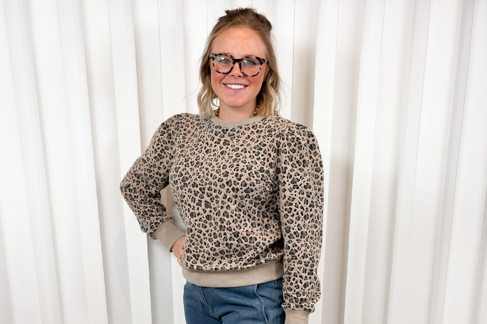 Woman wearing a leopard print sweater and blue jeans against a white curtain background